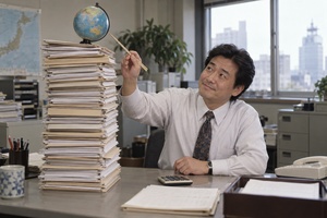 日本人の会計担当者が書類の山に乗った地球儀を箸で慎重に支える、海外子会社と資本のバランスを表す写真